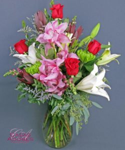 boldly blooming statement of red roses, cymbidium orchids, white oriental lilies, burgundy snapdragons and misty blue, overflowing from a modern vase. 