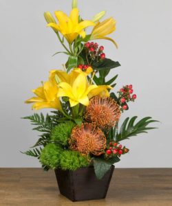 The design features bright yellow lilies, orange pincushion protea, red coffee beans and fun green dianthus styled in a modern, wood container