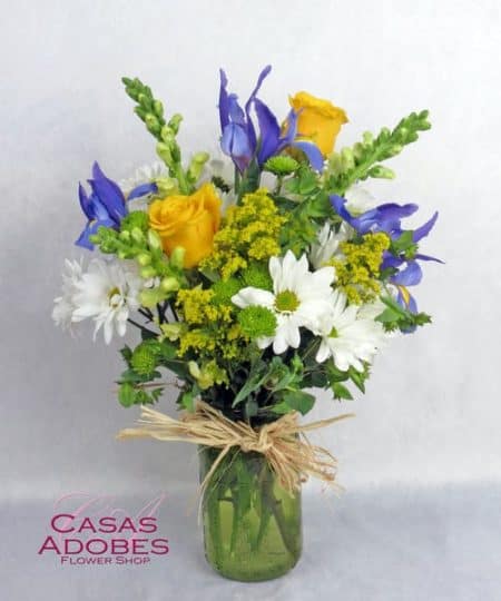 They will love this vibrant garden mix of blue iris, yellow roses and white daisy chrysanthemums accented with yellow solidago and green button chrysanthemums. This bouquet is artfully designed in a green mason jar shaped vase with a touch of raffia for a down home cottage feel.